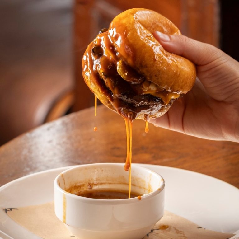 The Audley burger: Aged beef patty, double cheese, caramelised onions, pickled onions & gherkins, horseradish mayonnaise, and beef fat jus dipping sauce. The burger is being dipped into the sauce.